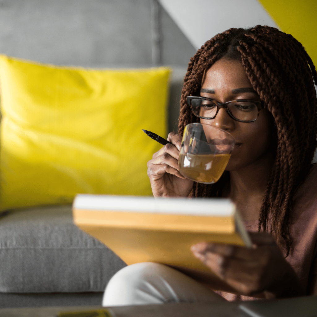A black woman sits on the floor in front of her couch with a notepad in front of her, and a pen and tea in her hand