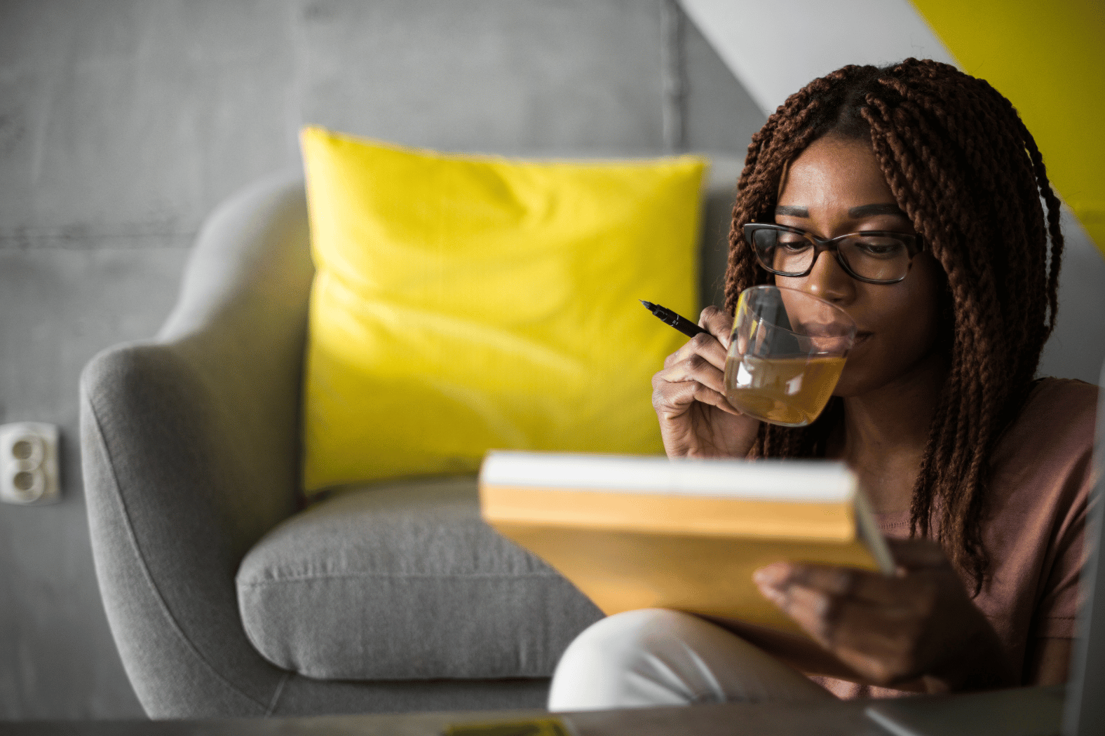 A black woman sits on the floor in front of her couch with a notepad in front of her, and a pen and tea in her hand