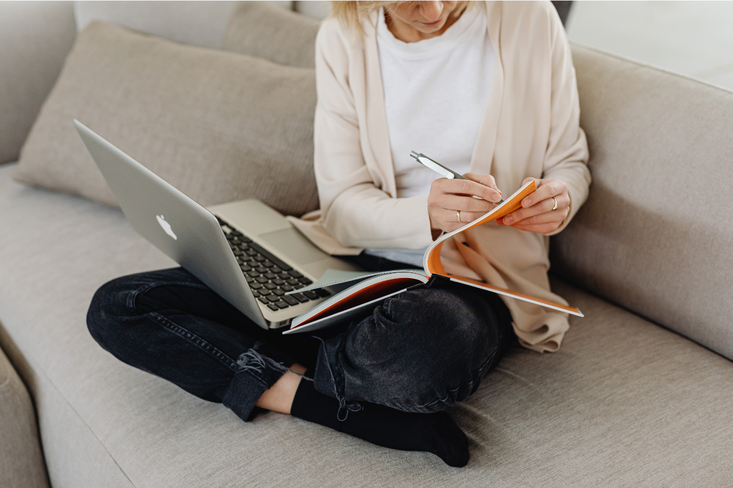Woman sits cross-legged on her couch with a notebook and laptop on her lap.