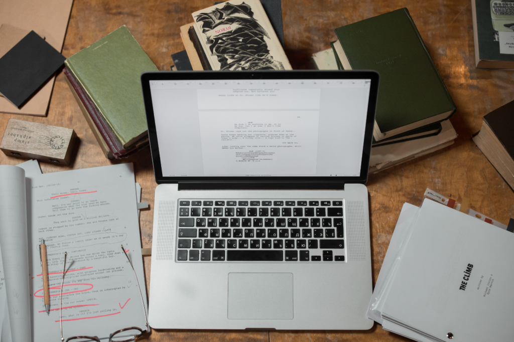 Laptop sides on a table covered with books, pens, and notebooks